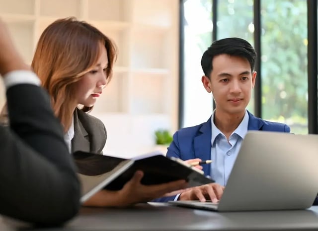 Two business professionals in formal attire reviewing documents and working on a laptop during a collaborative meeting.