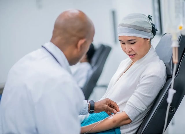 Clinician adjusts an IV catheter for a seated patient wearing a headscarf in an infusion clinic.