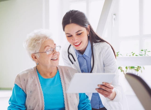A smiling doctor showing a tablet to an elderly woman, discussing health information in a bright medical setting.