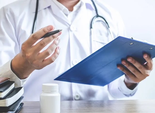 Doctor in a white coat with a stethoscope reviews a blue clipboard, pen in hand, with a white pill bottle on the desk.