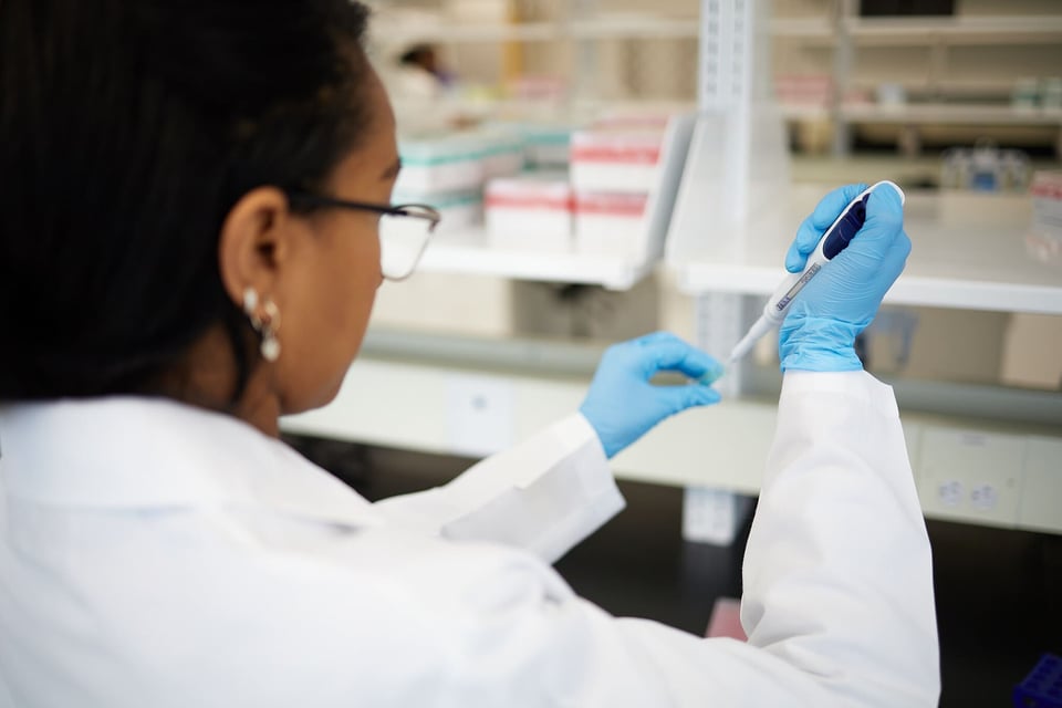 A scientist in a lab coat and blue gloves using a pipette to transfer liquid samples during customized assay preparation in a laboratory setting.
