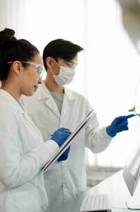 Two scientists in lab coats and safety glasses review data on a tablet in a lab setting, representing regulatory strategy, authoring, and submission support for IND, CTA, BLA, and MAA.